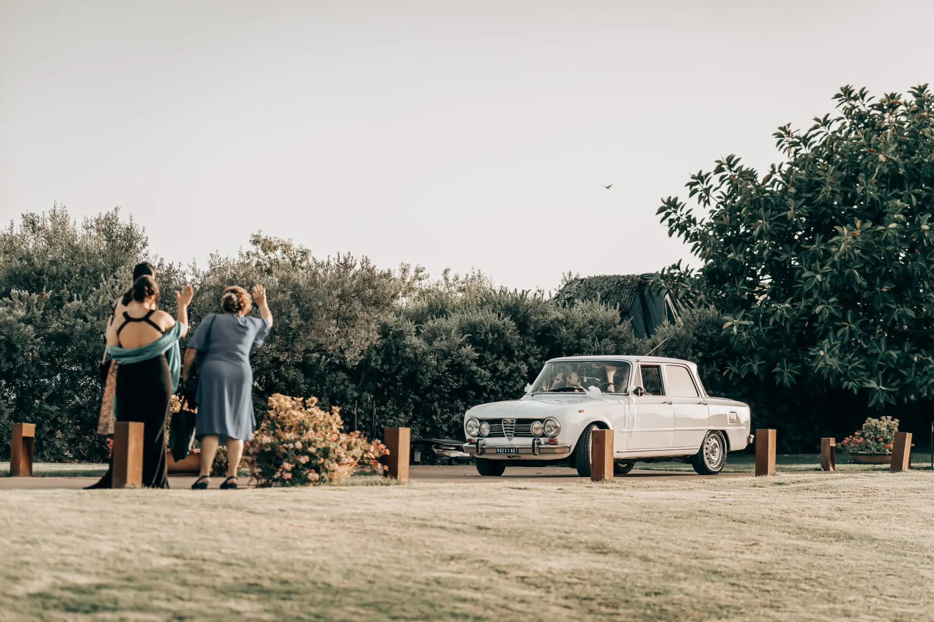 Scatto spontaneo di tre invitate ad un matrimonio all'aperto in Sicilia. La presenza dell'auto d'epoca bianca rafforza il tema vintage-elegante della cerimonia. Esempio di reportage fotografico di matrimonio che valorizza dettagli e momenti sinceri, realizzato dal fotografo Daniele Pullara.
